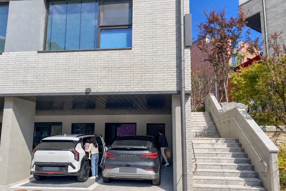 집 앞 주차 자리에서 엄마가 아이를 안아 내리고 아빠가 장바구니를 들고 현관으로 가는 연출 장면